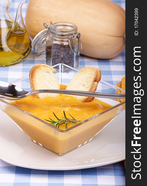 Cream of squash soup in a squared glass plate with toasted bread and rosemary. Pumpkin, grain pepper and olive oil in the background. Studio shot. Selective focus, shallow DOF.