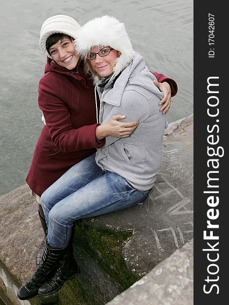 Outdoor shot of two young, pretty woman with white funny woolen caps at the lake, friendly smiling and embracing. Outdoor shot of two young, pretty woman with white funny woolen caps at the lake, friendly smiling and embracing