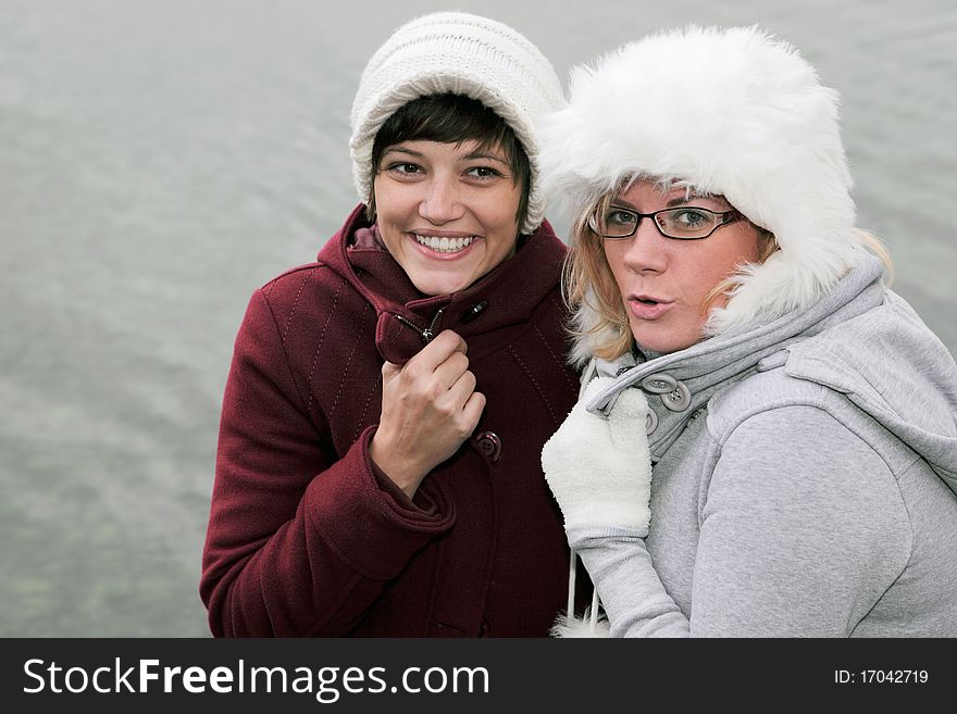 Winter outdoor shot at a lake of two young, pretty woman in winter clothing with white woolen hats, friendly smiling into the camera. Winter outdoor shot at a lake of two young, pretty woman in winter clothing with white woolen hats, friendly smiling into the camera.