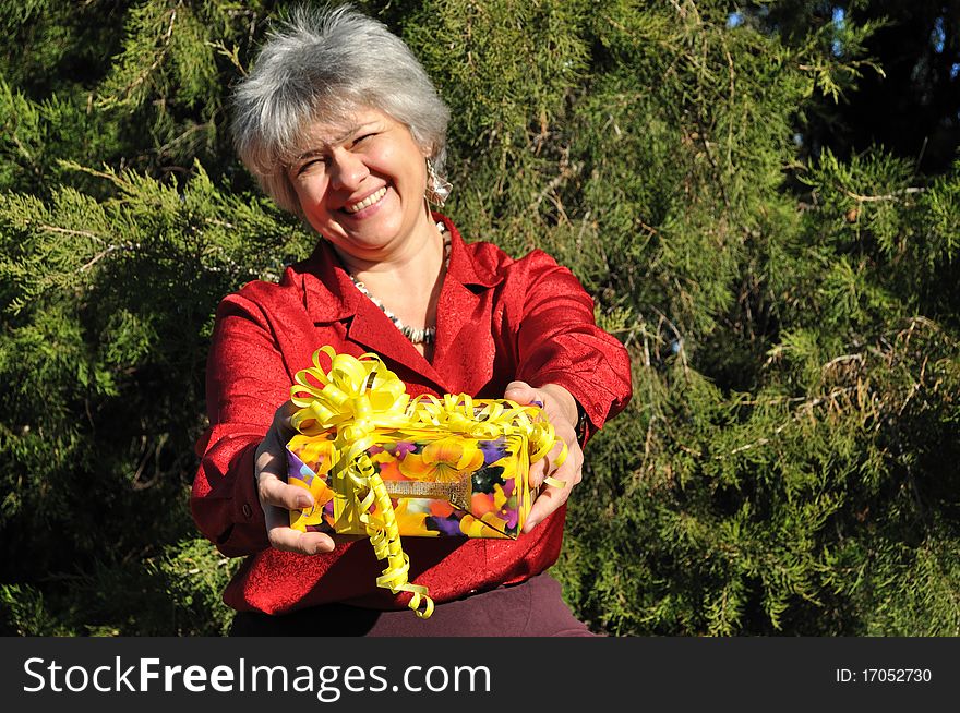Lucky, an old woman holds out a gift in the yellow box, wrapped in gift paper and ribbons, against the backdrop of trees. Lucky, an old woman holds out a gift in the yellow box, wrapped in gift paper and ribbons, against the backdrop of trees