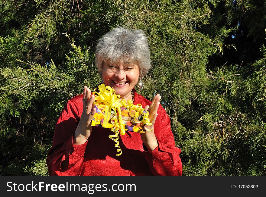 A happy, middle-aged woman holding a gift with both hands, against the backdrop of trees