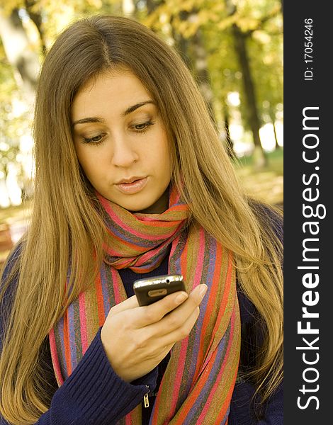 Young Caucasian woman calls on a mobile phone, sitting in a park on a wooden bench. Autumn around a lot of colorful foliage. Surprised. SMS. Young Caucasian woman calls on a mobile phone, sitting in a park on a wooden bench. Autumn around a lot of colorful foliage. Surprised. SMS