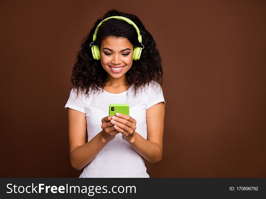 Portrait of positive cheerful afro american girl use cellphone want listen, radio melody search online playlist have green wireless headset isolated over brown color background. Portrait of positive cheerful afro american girl use cellphone want listen, radio melody search online playlist have green wireless headset isolated over brown color background