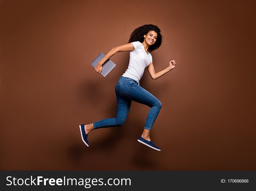 Full length profile photo of funny dark skin lady jumping high holding, notebook hurry classes lessons schoolgirl wear casual white t-shirt jeans isolated brown color background. Full length profile photo of funny dark skin lady jumping high holding, notebook hurry classes lessons schoolgirl wear casual white t-shirt jeans isolated brown color background