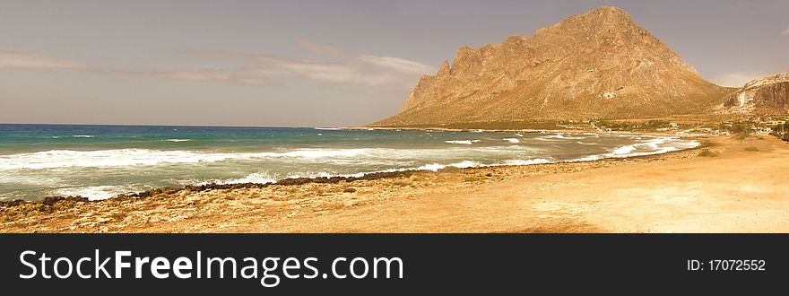 Long horizontal seaside panorama of a bay; rocky beach, sandy beach, resort village and hill in the distance Location: Cornino village, Gulf of Bonagia and view of Monte Cofano mountain in Sicily, Italy, Europe 3 horizontal images stitched together into a panoramic image