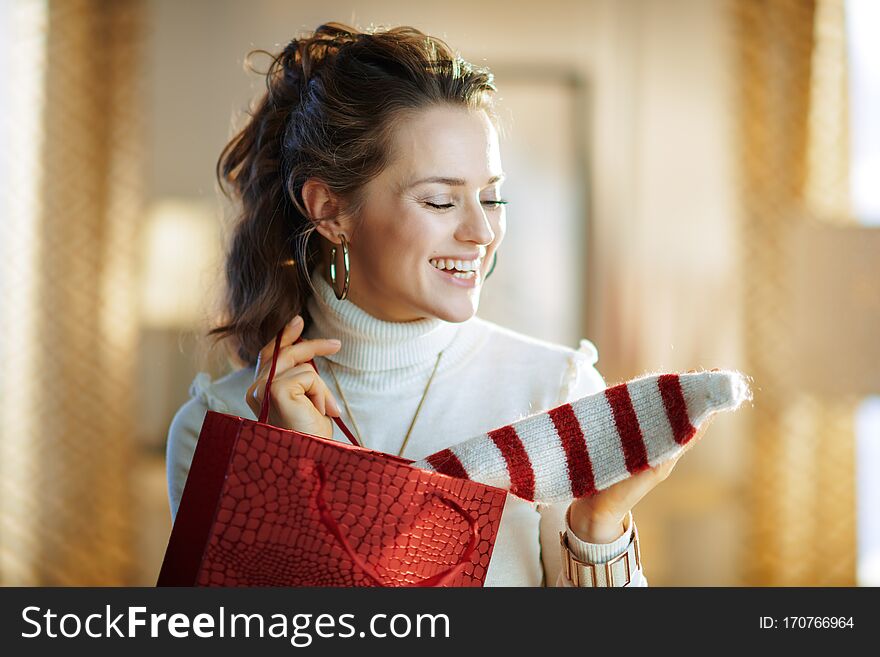 Happy stylish woman in white sweater and skirt with red shopping bag taking out purchased sweater in the modern house in sunny winter day. Happy stylish woman in white sweater and skirt with red shopping bag taking out purchased sweater in the modern house in sunny winter day