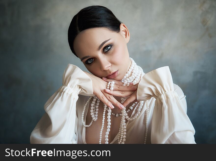 Girl dressed in white boho clothing with white pearl beads around her neck is sitting at a table. Perfect smile, romantic sexy image of a woman, clean smooth skin and beautiful makeup