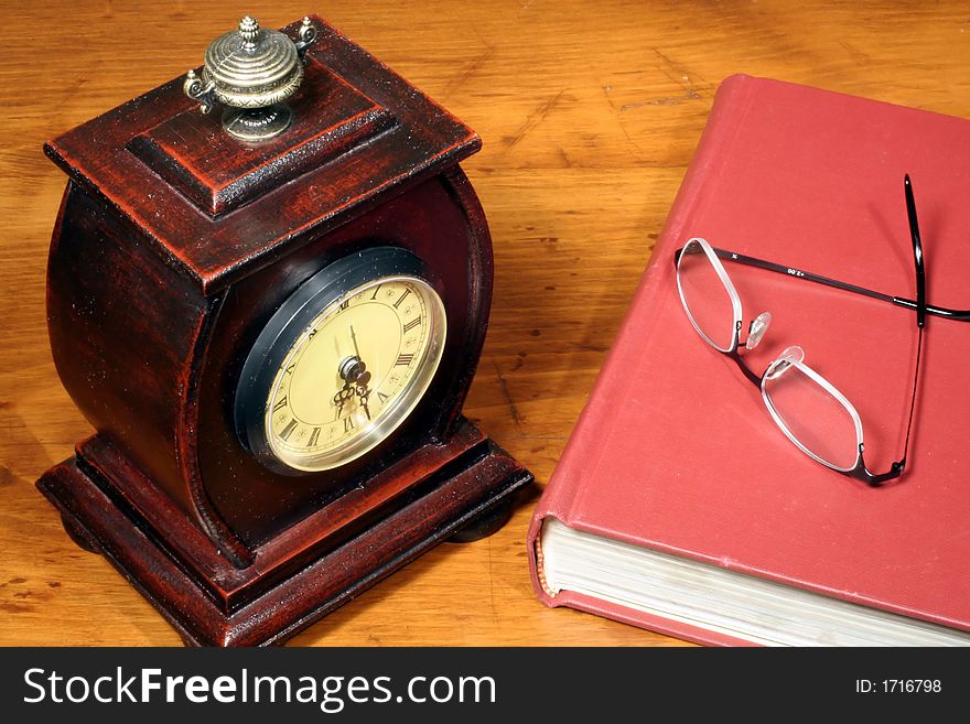 This is an image of a clock, glasses, and book on a wooden desktop. This is an image of a clock, glasses, and book on a wooden desktop.