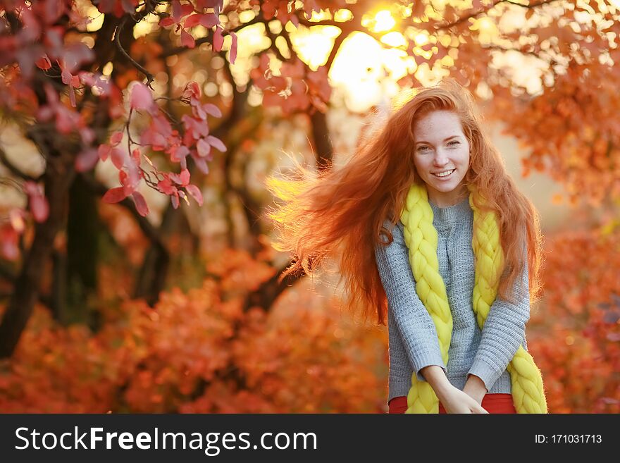 A woman stands in a forest with falling red leaves and looks directly at the camera.