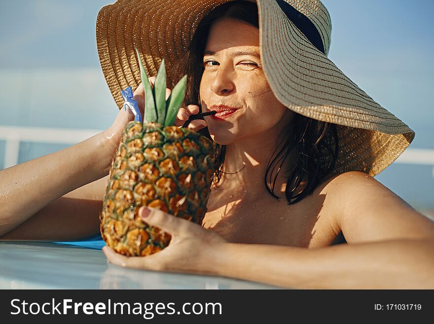 Beautiful young woman smiling and drinking cocktail in pineapple, relaxing in pool on summer vacation. Girl in hat enjoying warm