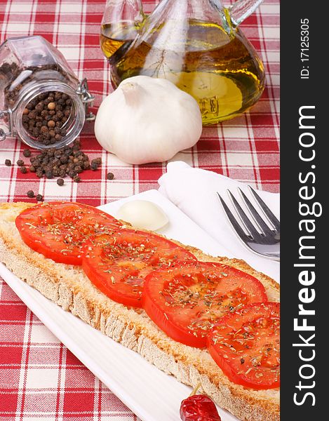 Toasted bread topped with tomato, olive oil and oregano served over a white plate with a garlic clove and a small red chilli pepper. Pepper, garlic and olive oil in the background. Toasted bread topped with tomato, olive oil and oregano served over a white plate with a garlic clove and a small red chilli pepper. Pepper, garlic and olive oil in the background.