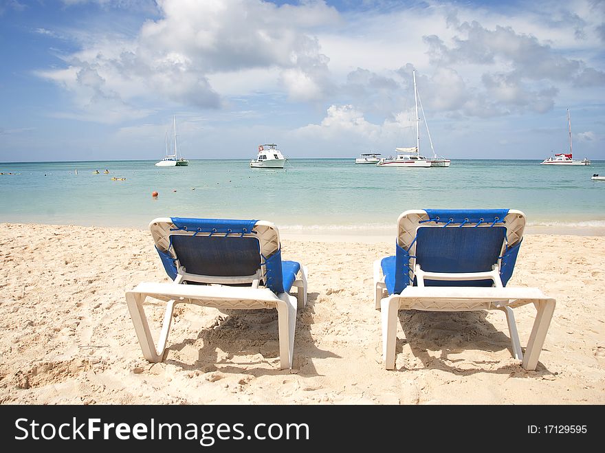 Empty blue beach chairs on a sandy beach overlooking the Caribbean Sea on a sunny day. Sailboats anchored in the background. Empty blue beach chairs on a sandy beach overlooking the Caribbean Sea on a sunny day. Sailboats anchored in the background.