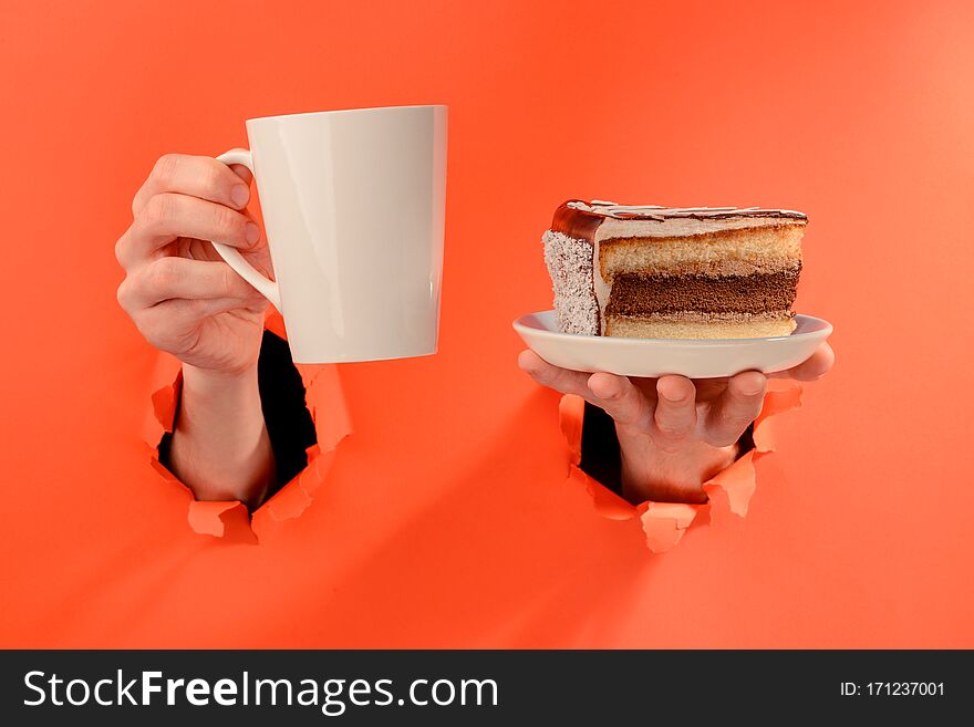 Hand holding a mug of coffee and a piece of chocolate cake through torn holes in red paper background.