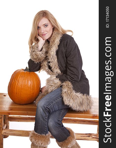 A woman sitting on a bench in her warm fuzzy coat while she leans on a pumpkin. A woman sitting on a bench in her warm fuzzy coat while she leans on a pumpkin.