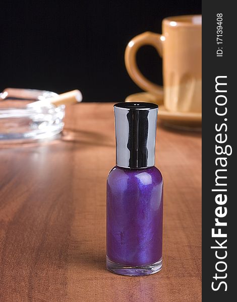 A bottle of purple nail polish standing on a coffee table in front of a cup of coffee and an ashtray.