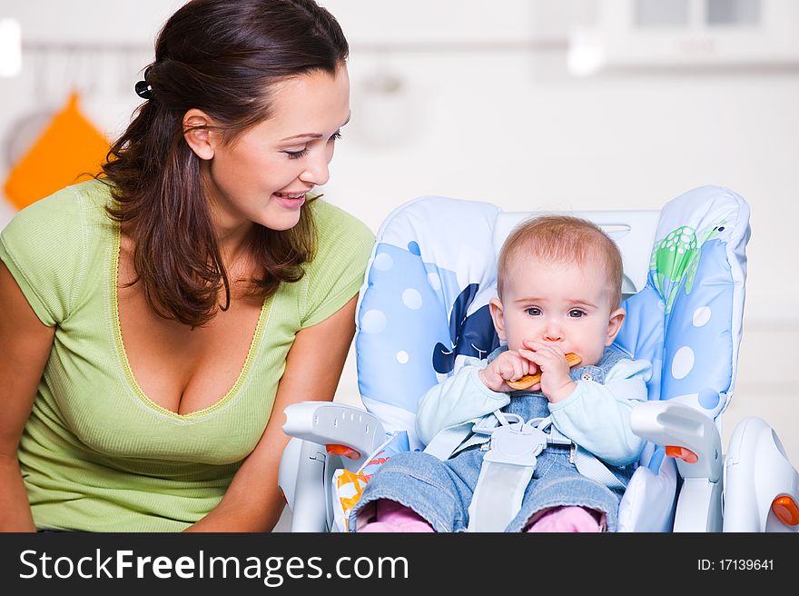 Happy mother looking on baby in the highchair. Toddler eating the cookie. Happy mother looking on baby in the highchair. Toddler eating the cookie