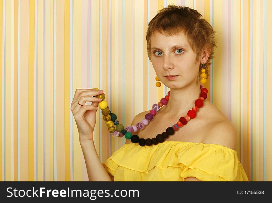 Young woman against yellow striped walls in a yellow blouse with bare shoulders in her wearing a necklace and earrings made of wool. Ethnos