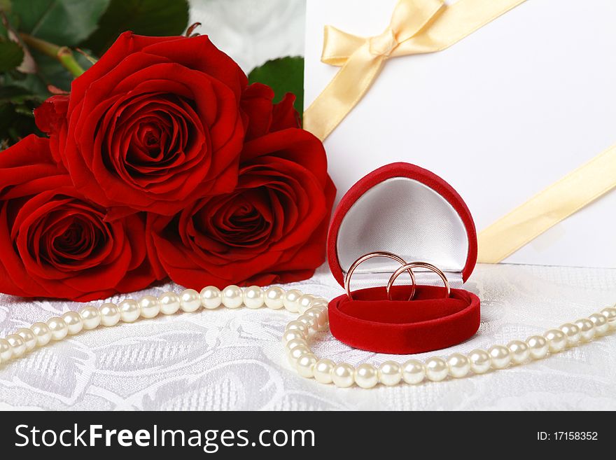 Wedding rings in box against bouquet of red roses and invitation card on lace background