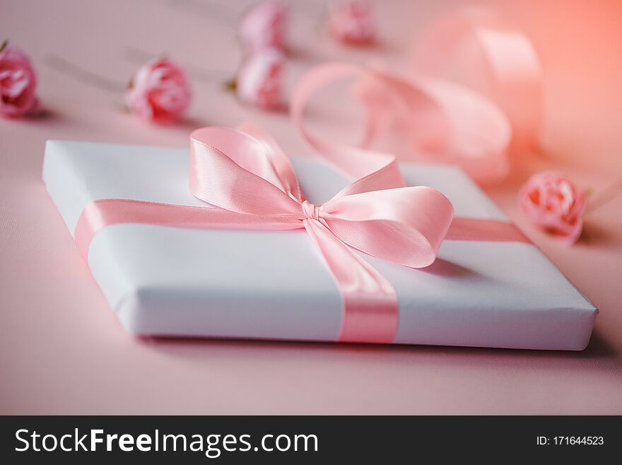 Composition of a white box and small pink flowers on a light pink background. Great beautiful pink ribbon bow. Packing of gifts. Composition of a white box and small pink flowers on a light pink background. Great beautiful pink ribbon bow. Packing of gifts