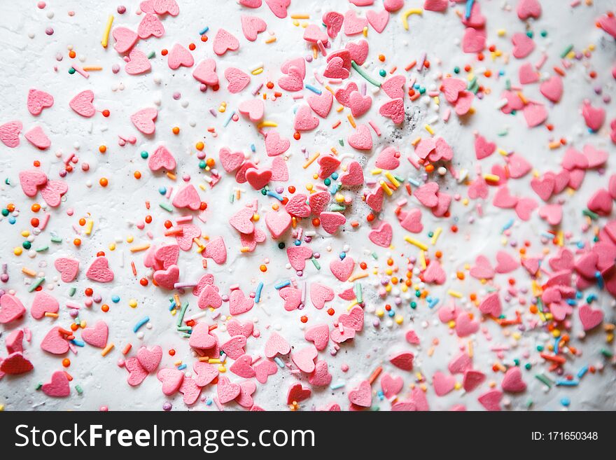 On a white sugar backing lies a sweet pastry topping in the form of pink hearts, orange, turquoise and red peas, and green, blue and yellow sticks