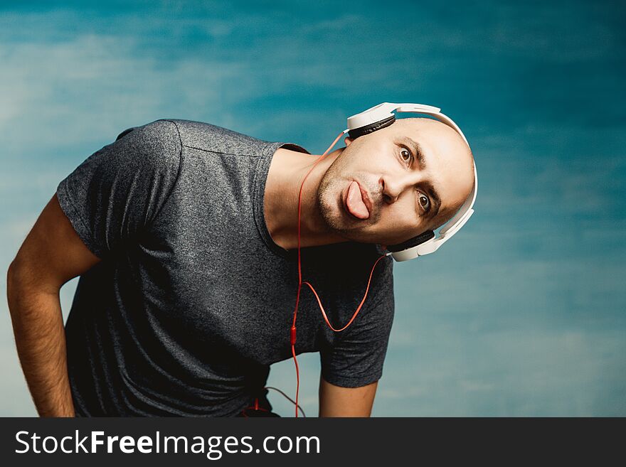 An interesting bald man in headphones is listening to music and leaning towards the camera shows his tongue on a blue