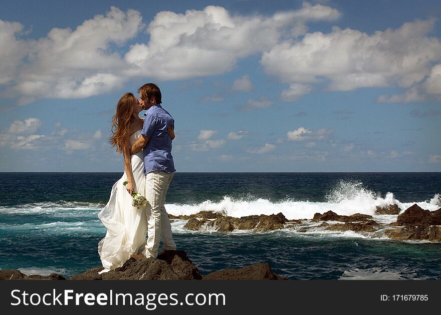 Young just married couple kissing on ocean rock shore. Side view. Ocean waves, splash of water and blue cloudy sky.