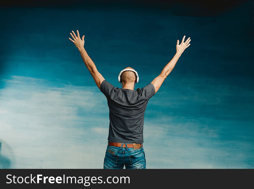 A Bald Man In Headphones Listens To Music, Standing With His Back To The Camera, Raising His Hands Up On A Blue