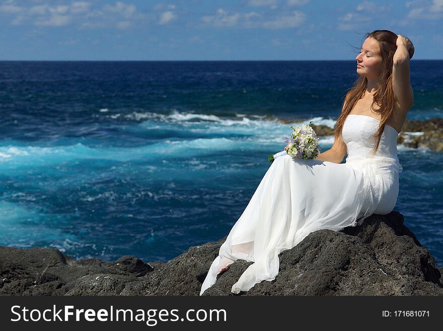 Elegant bride in light white wedding dress sitting on ocean rock shore, flowers in hand. Wedding ceremony on ocean coast