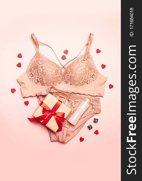 Vertical photo of lace women`s pink underwear, gift box with red ribbon, lubricant, cubes and heart shape on a pink background