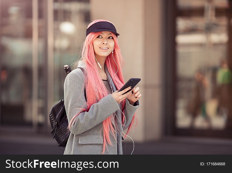 Japan anime cosplay. Colorful portrait of a young attractive smiling Asian woman in a coat with creative make-up wearing a pink wig. Trendy Japanese girl using smartphone listening music in earphones walking on the city street. Japan anime cosplay. Colorful portrait of a young attractive smiling Asian woman in a coat with creative make-up wearing a pink wig. Trendy Japanese girl using smartphone listening music in earphones walking on the city street.