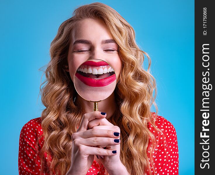 Blonde holds a magnifying glass in front of her teeth. Girl with a bright smile. Funny girl. Young woman holds a magnifying glass in front of her teeth. More teeth. Dental care. Funny face of a girl