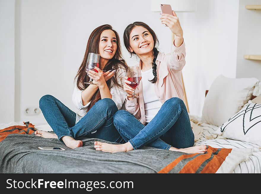 Young carefree brunette girls friends in casual with glasses of wine taking selfie and having fun together on home party