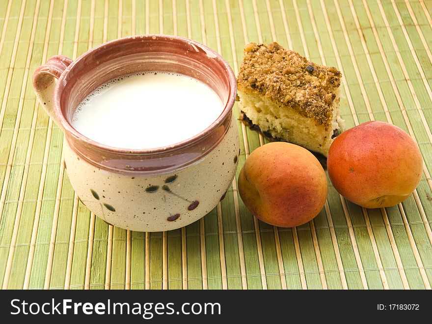 A cup of milk, plate with cake and a ripe apricot on the green mat. A cup of milk, plate with cake and a ripe apricot on the green mat