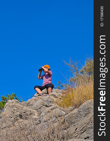 Boy sitting on a rock on top of a mountain and sees the neighborhood through binoculars. Boy sitting on a rock on top of a mountain and sees the neighborhood through binoculars