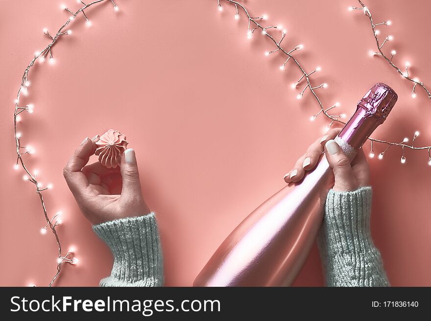 Valentine top view on pink background. Light garland, woman hands with metallic pink champagne bottle and sweet marshmallow snack