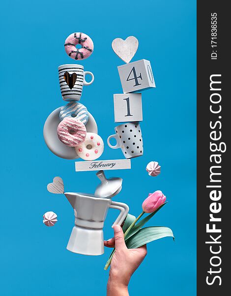 St. Valentine`s day fun background in blue mint . Date 14 February on wooden calendar with coffee cups, tulip, pink donuts and marshmallows. Female hand holding balancing pyramid on coffee maker