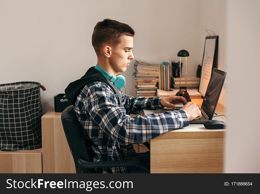 Teenage boy doing homework using computer sitting by desk in room alone. Teenager warching video and browsing internet on personal laptop