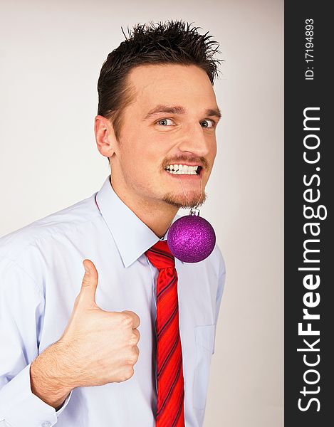 Happy young man in shirt and tie has violet Christmas Ornament between teeth and shows with thumb yes. Happy young man in shirt and tie has violet Christmas Ornament between teeth and shows with thumb yes.