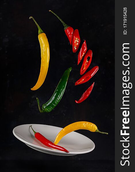 Flying red, yellow and green chili peppers on a dark background with a ceramic plate. Flying red, yellow and green chili peppers on a dark background with a ceramic plate