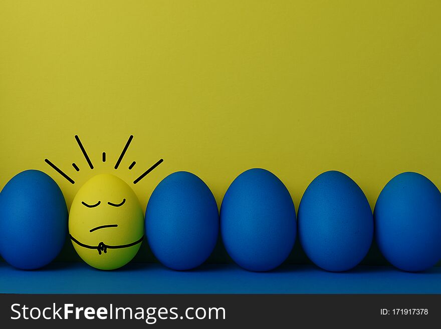 Five blue and one yellow funny faced painted Easter eggs stand on a yellow with blue background. Copy space. Creative Easter concept. Five blue and one yellow funny faced painted Easter eggs stand on a yellow with blue background. Copy space. Creative Easter concept