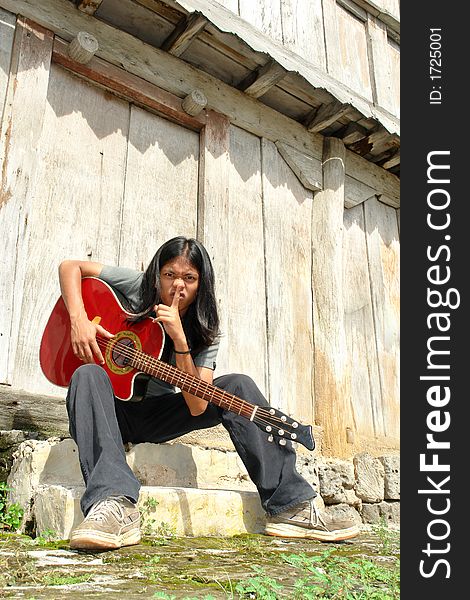 Asian exotic long-haired boy sitting in front of an old wooden building with an acoustic guitar, explicitly asking for silence. Asian exotic long-haired boy sitting in front of an old wooden building with an acoustic guitar, explicitly asking for silence.