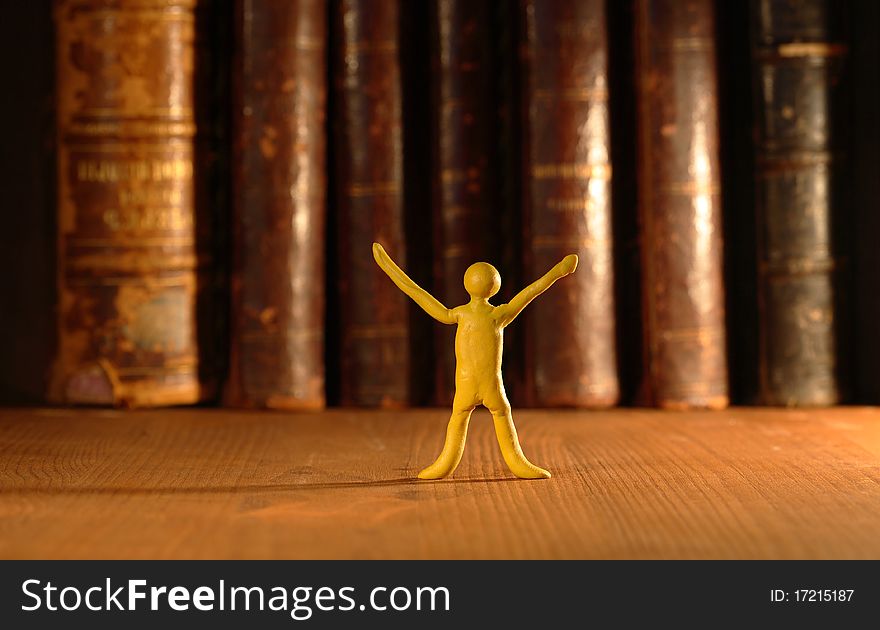 Small yellow plasticine man standing on background with old books in a row. Small yellow plasticine man standing on background with old books in a row