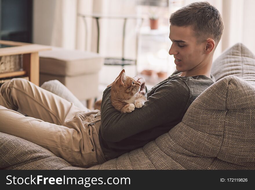 Owner playing with cat while relaxing on modern couch in living room interior. Young man resting with pet in soft chair at home. Owner playing with cat while relaxing on modern couch in living room interior. Young man resting with pet in soft chair at home