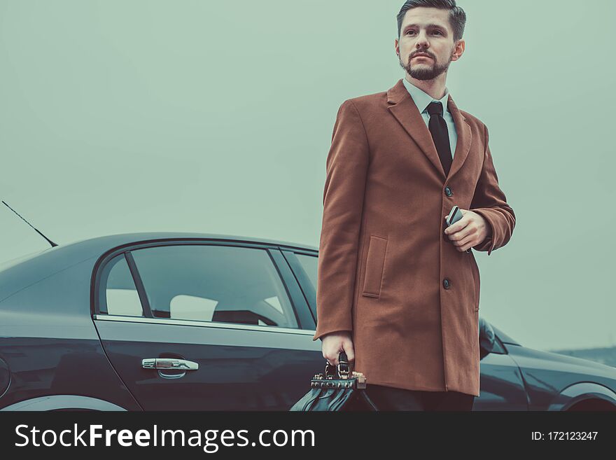 Portrait of a young businessman, in a business style, holding a leather bag in his hands, stands by the car. In creative