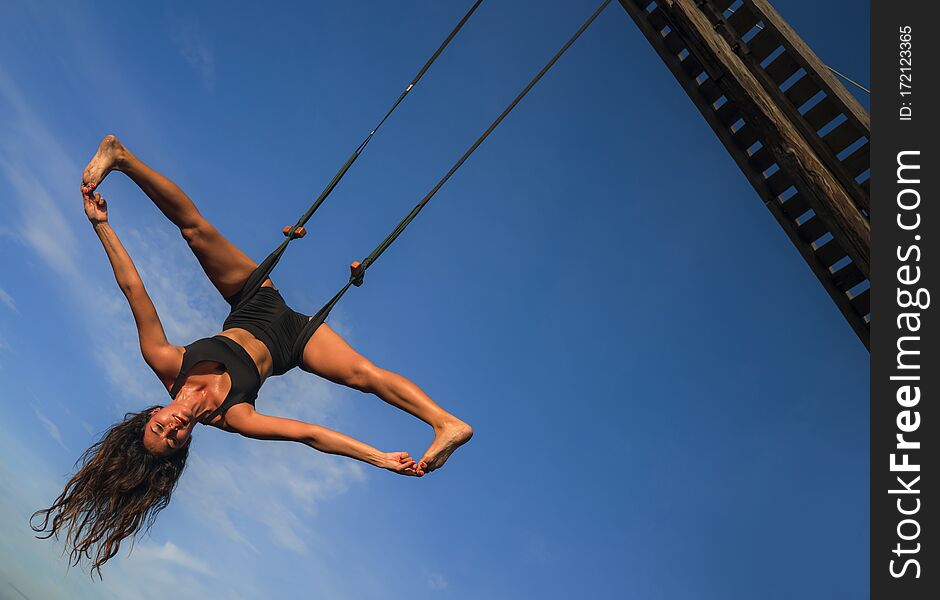 Aero yoga workout - young attractive and athletic woman practicing aerial yoga exercise training acrobatic  body postures isolated on blue sky in balance flexibility control and healthy lifestyle