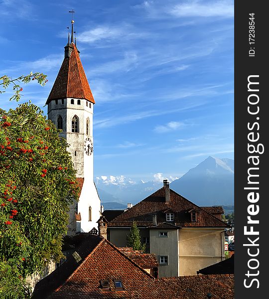 The clock tower with red tiled roof raising over the buildings of the old city, against the background of bright blue sky, with a view of distant mountains