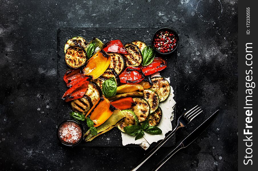 Grilled multicolored vegetables, aubergines, zucchini, pepper with green basil on serving stone board on black background, top