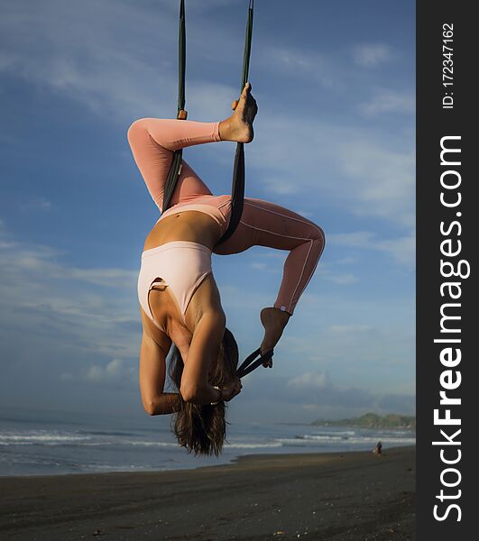 Aerial yoga beach workout - young attractive and healthy woman practicing aero-yoga training balance body and mind control hanging from ropes above the sea isolated on beautiful blue sky