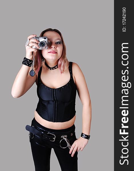 A young punk woman standing in black leather pants and a black corset with a camera in her hand for gray background. A young punk woman standing in black leather pants and a black corset with a camera in her hand for gray background.