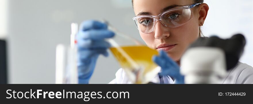 Close-up of laboratory researcher holding medical glass bottle with yellow liquid wearing sterile gloves and special lab eyewear. Scientist woman at job. Chemistry concept. Close-up of laboratory researcher holding medical glass bottle with yellow liquid wearing sterile gloves and special lab eyewear. Scientist woman at job. Chemistry concept